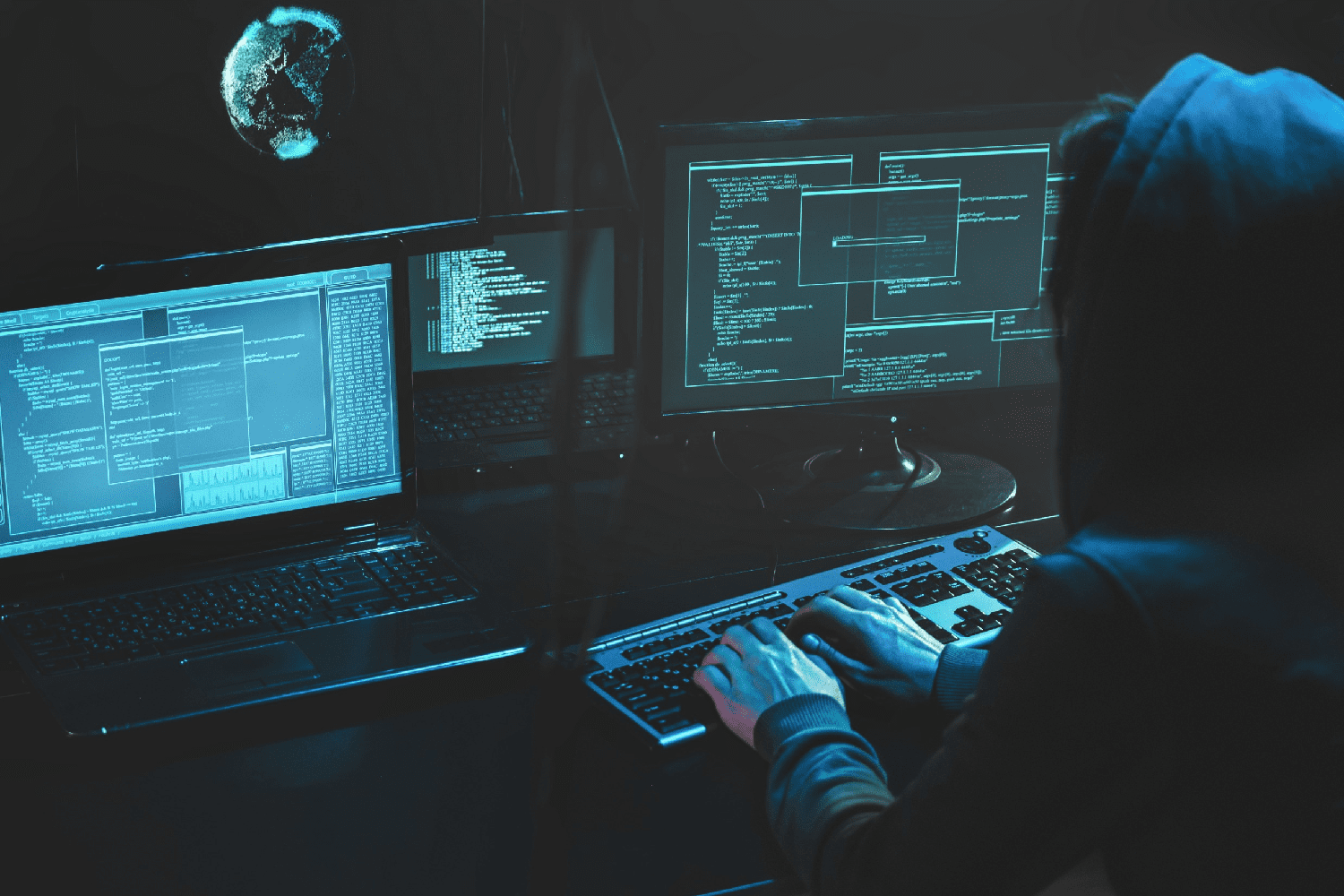 A cybercriminal in a hoodie is hacking a computer and attacking web servers in a dark room.